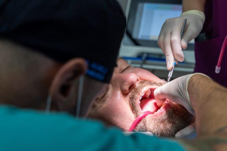 The dentist and dental assistant work together to place a tooth-shaped implant restoration into a prepared socket while treating a male patient.